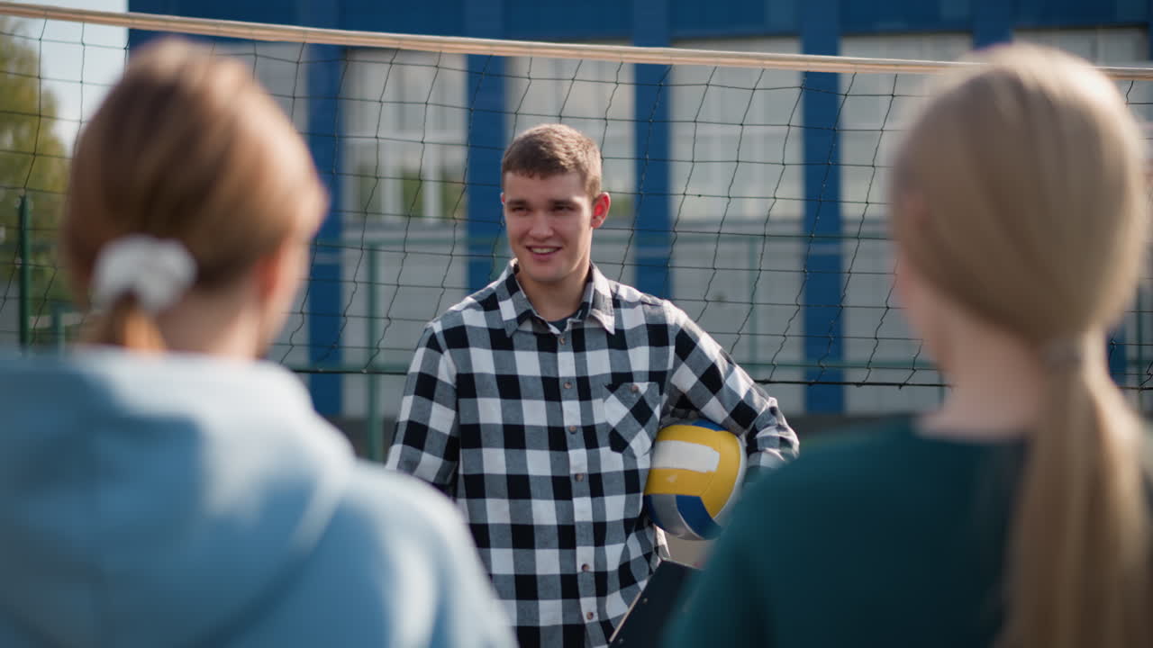 배구 코트에서 새로운 학생을 가르치는 동안 미소 짓는 젊은 코치, 배경에는 네트, 코트, 그리고 뒤에 학생들의 흐릿한 건물이 있습니다.
