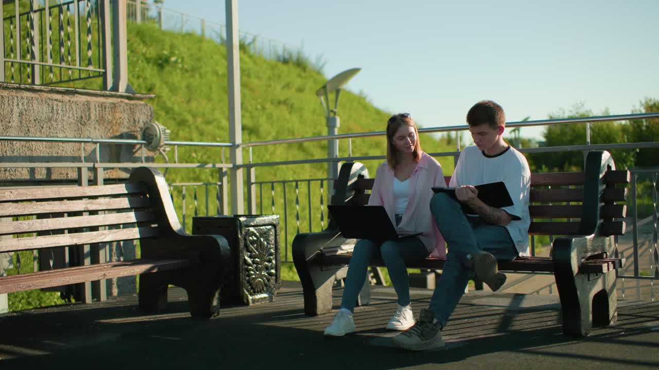 Woman in pink seated outdoors studying with friend, she withdraws hand from his tablet, continues on her laptop, with green hill and light poles in the background