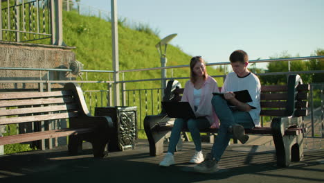 woman in pink seated outdoors studying with friend, she withdraws hand from his tablet, continues on her laptop, with green hill and light poles in the background