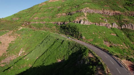 Aerial-view-of-car-driving-on-mountain-road-in-a-lush-sunny-green-landscape