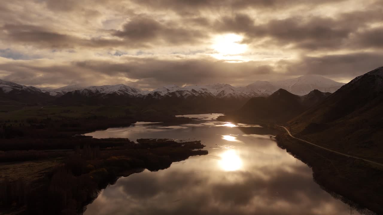 Sunset reflection over the water of Waitaki river near snowy mountains in background, aerial shot