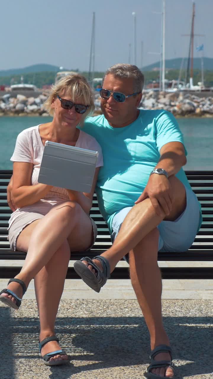 Pareja de Mediana Edad Sentada en Banco Junto a la Marina y Usando Tableta
