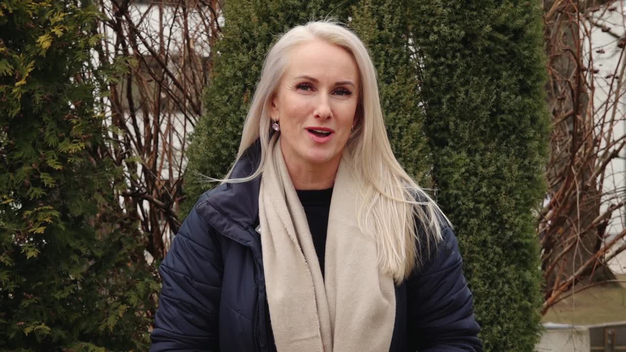 A woman with long blonde hair smiles and speaks while standing outdoors in front of evergreen trees, dressed warmly in a navy coat and beige scarf.