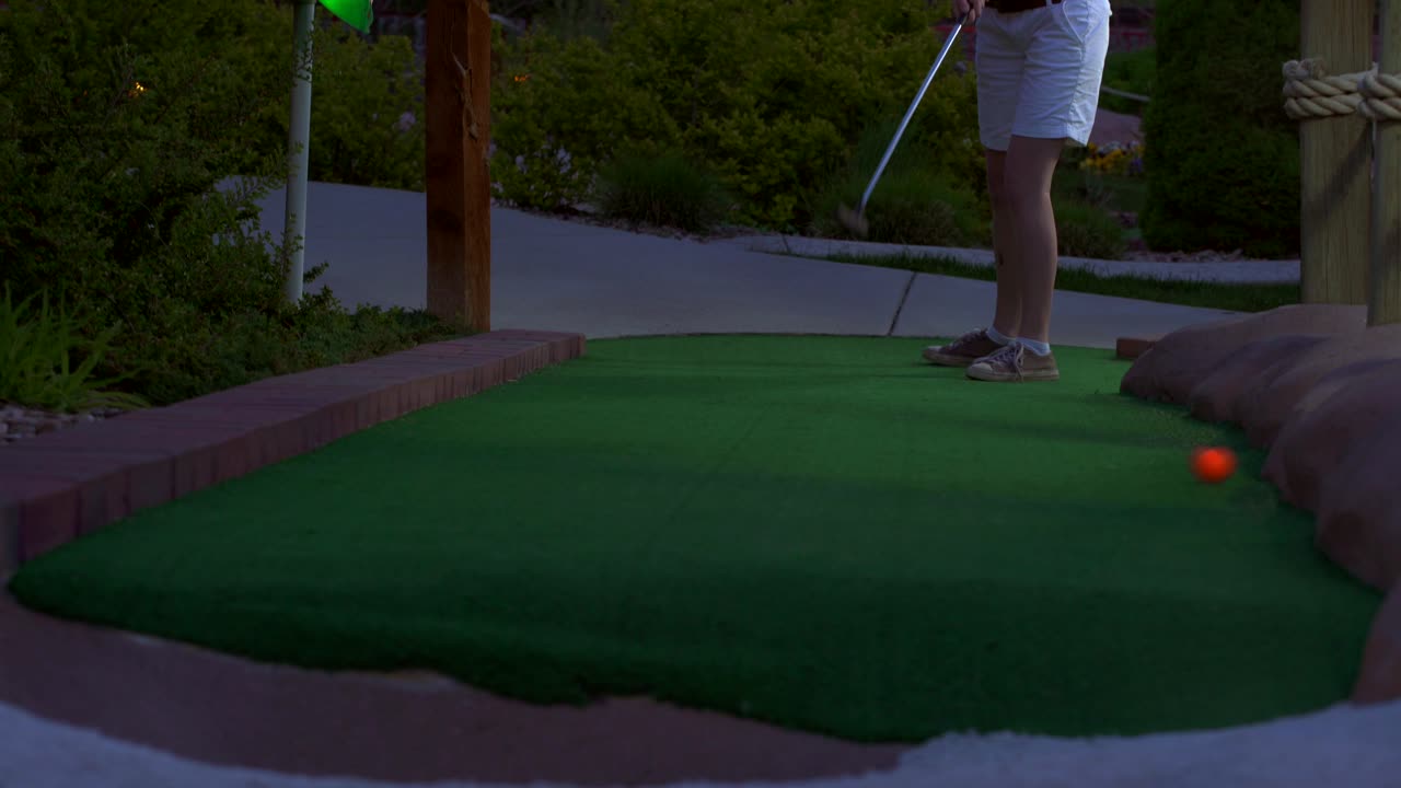 A caucasian girl mini golfing an orange golf ball hitting it towards the camera