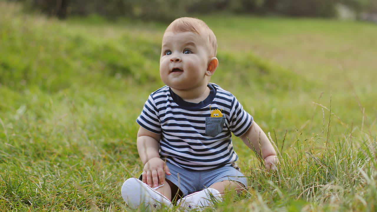 Portrait Of A Lovely Baby Boy Sitting On The Green Grass And Smiling In The Park