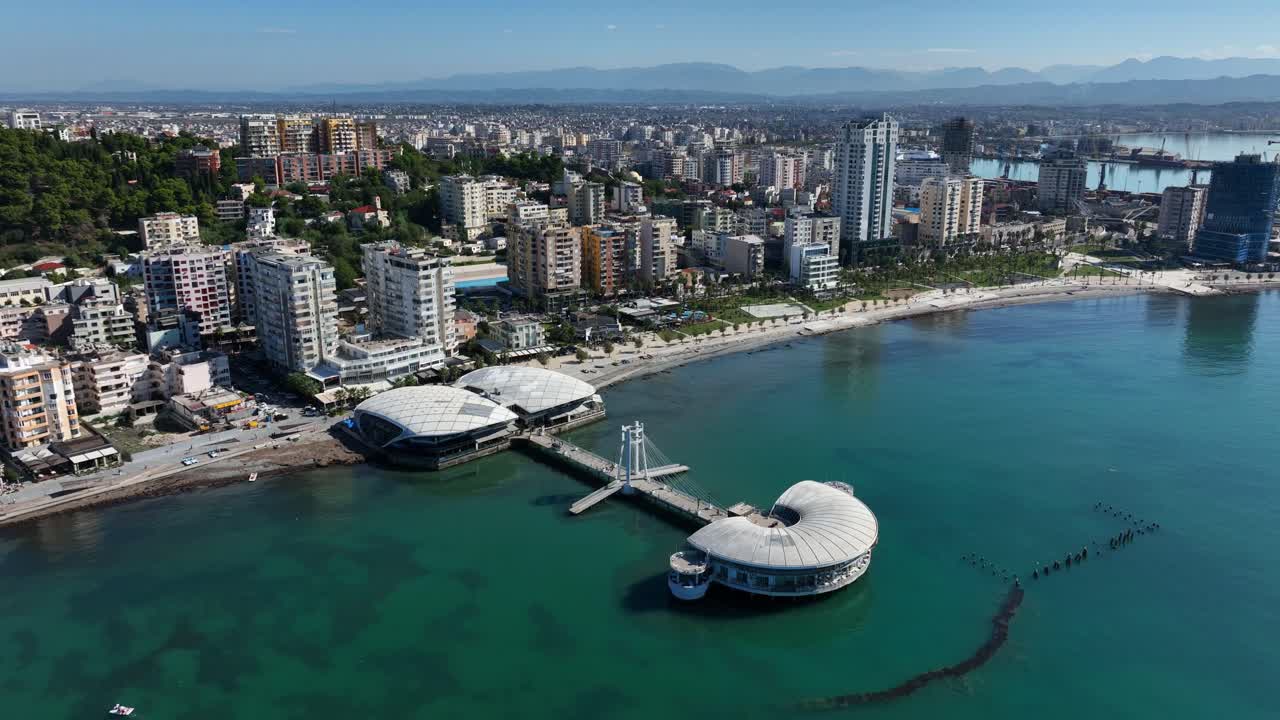 Aerial orbit tilting down across of Durres pier and surrounding urban coastline on a calm sunny day in Albania