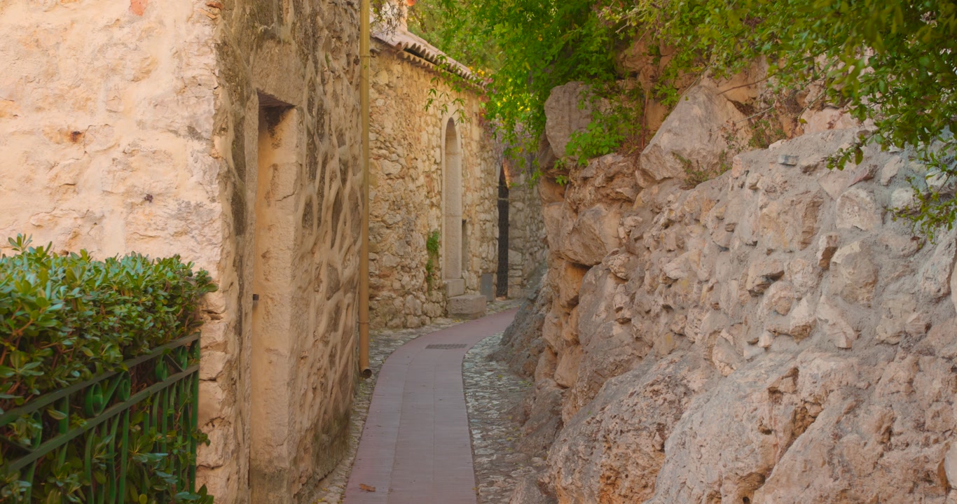 Narrow cobblestone alleyway winds through ancient stone walls of Eze, a perched medieval village on the French Riviera. Lush greenery contrasts with golden limestone architecture.