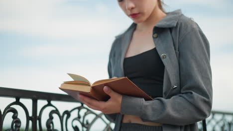 lady in open grey shirt holding an open book with right hand on pages and left hand beneath, reading with focus, wind softly blows the book's pages, blurred background shows water body