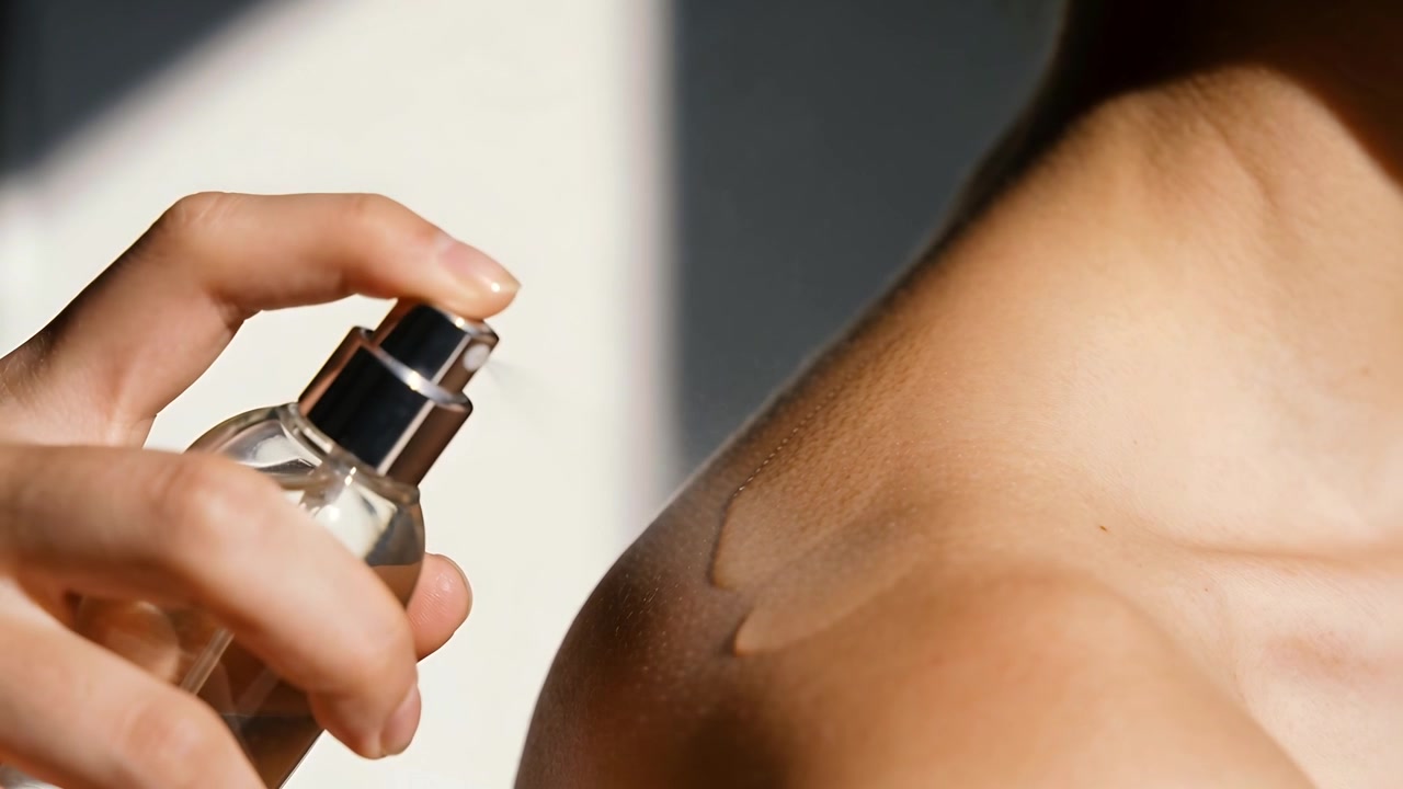 Mano che preme un flacone spray a pompa vicino a un petto nudo in studio, la luce crea goccioline che formano una scia lucida.