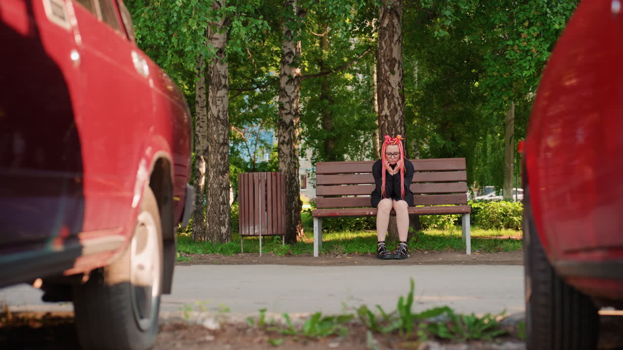 Белая женщина с неоновыми косами ждет на скамейке у дороги, красные автомобильные колеса обрамляют передний план, перспектива тихой улицы, березы и тротуар на заднем плане, ощущение дистанции и напряжения, откровенный городской пейзаж