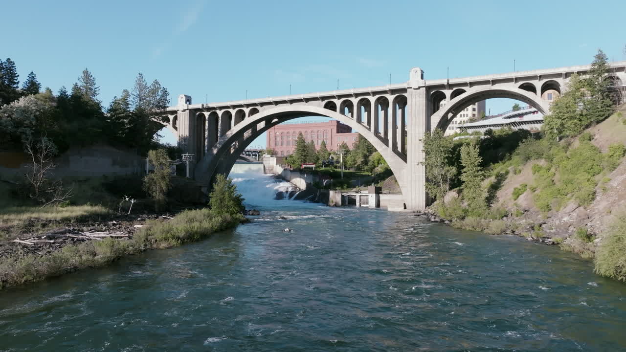 A downstream view of the Spokane River rushing beneath the Monroe Street Bridge with Spokane’s redbrick skyline and lush green banks on both sides