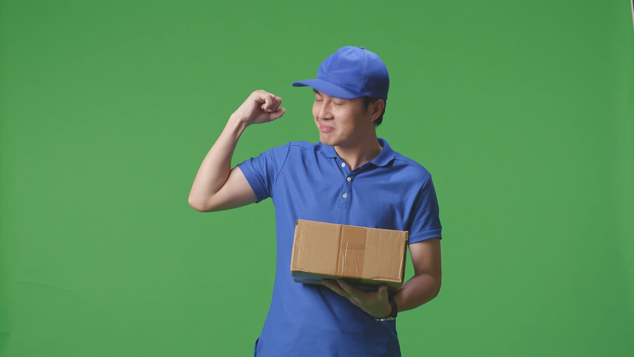 Asian Male Courier In Blue Uniform Smile And Flex Muscle While Delivering A Carton On Green Screen Background In The Studio