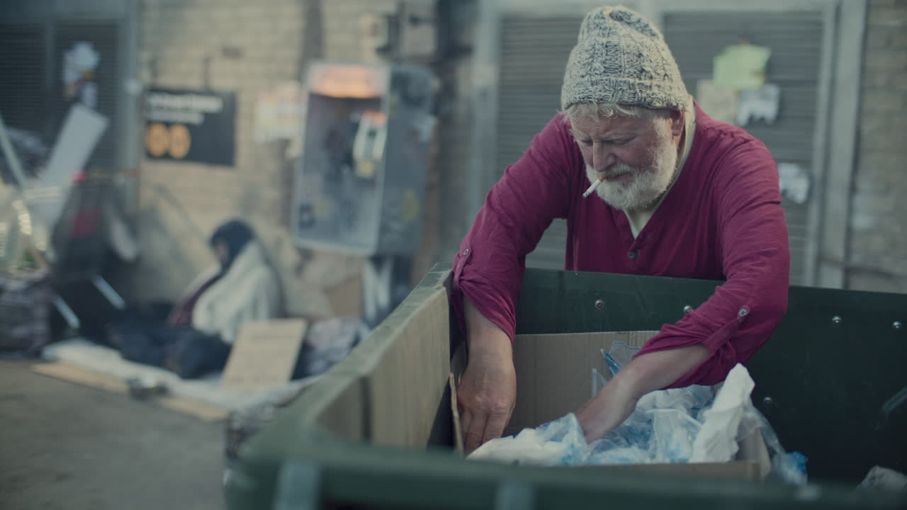 Elderly Homeless Man Smoking and Digging through Trash Bin on Street
