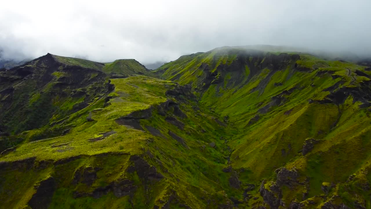 Aerial drone footage flying closer to tall green moss and nature covered Iceland volcanic landscape mountains during a cloudy day. Steep and rough valleys in between the mountains and clouds visible.