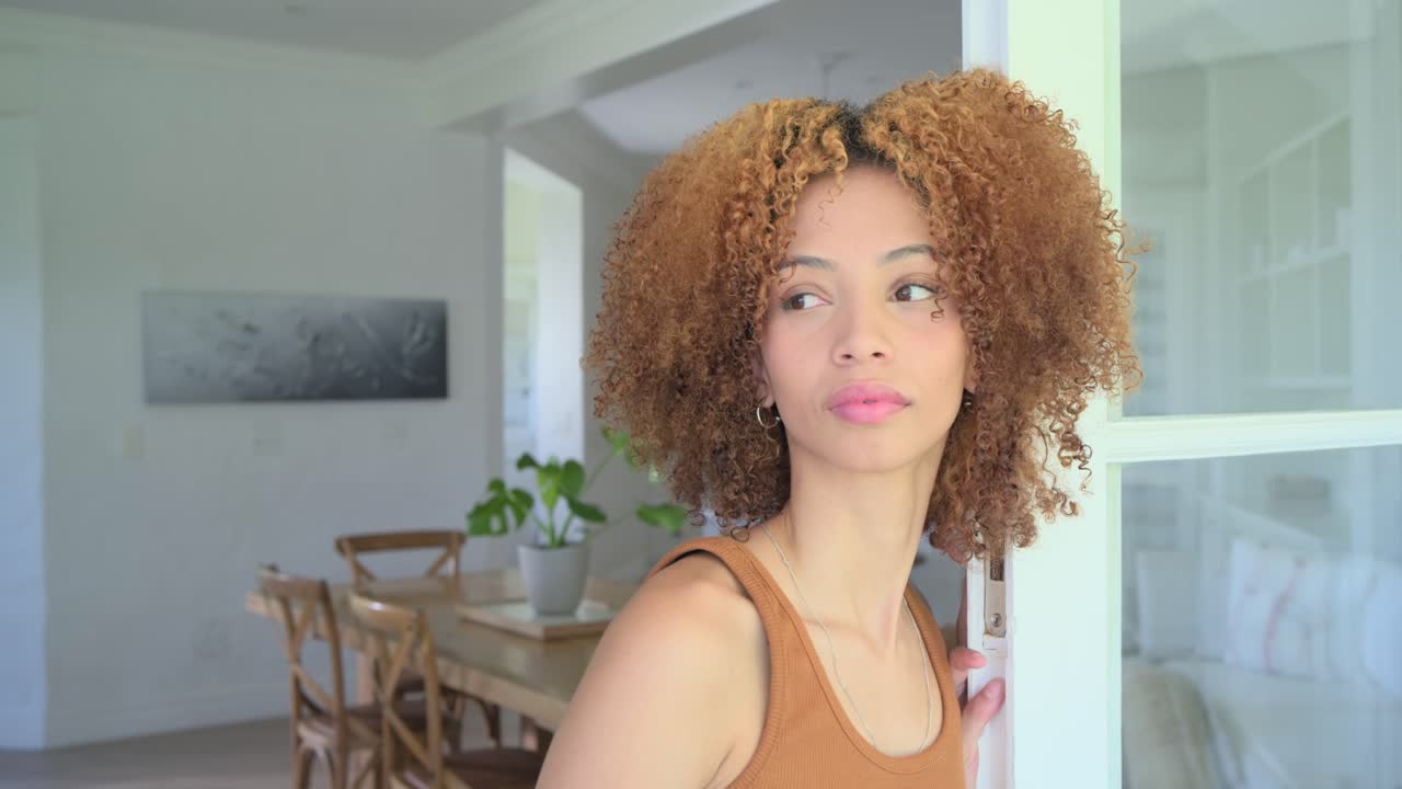 Mujer afroamericana apoyada en la puerta, escuchando una voz, girando la cabeza, cerrando los ojos y sonriendo, con una camiseta de tirantes.