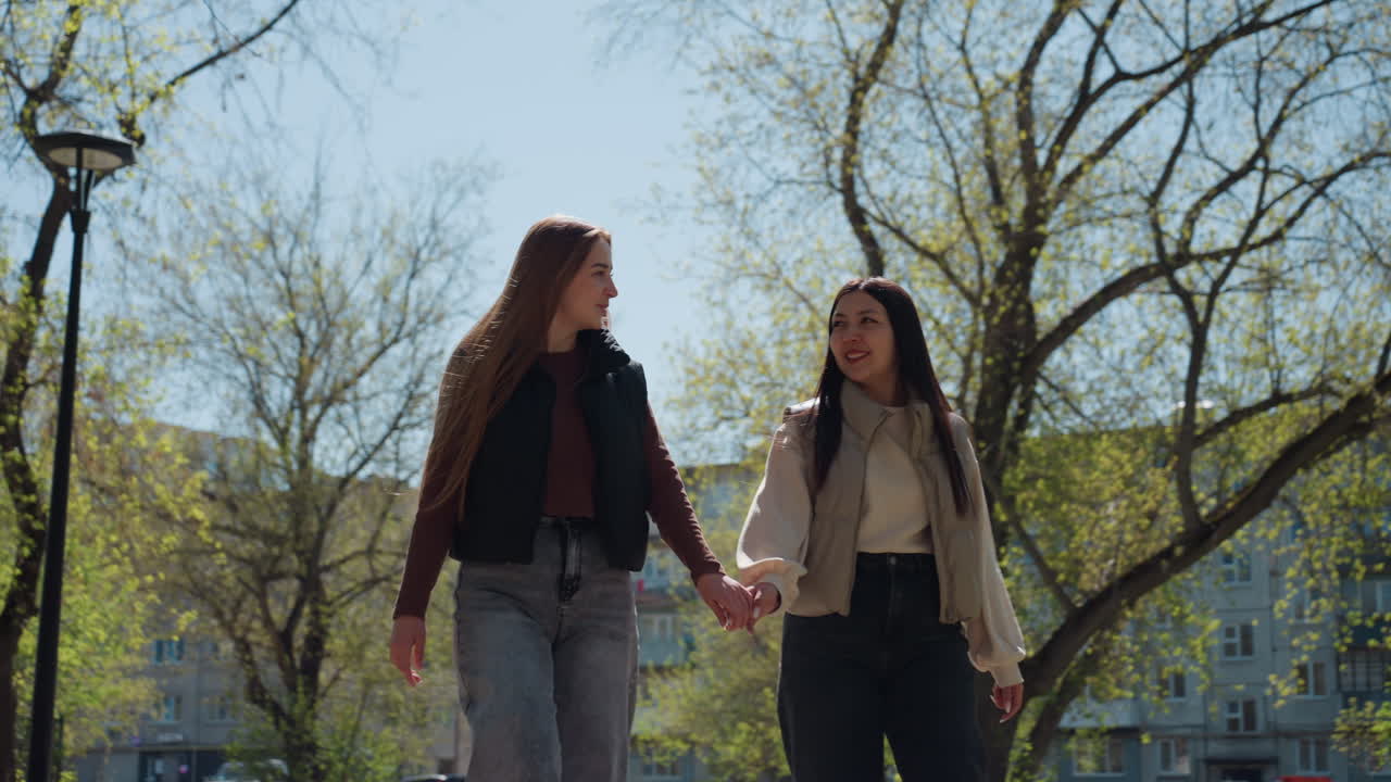 백인 여성과 아시아 여성이 가로등과 싹이 트는 나무를 지나 걸어가면서 손을 잡고 있고, 멀리 도시 스카이라인이 보이며, 차분한 속도와 사려 깊은 표정, 봄 오후, 도시 공원 산책로