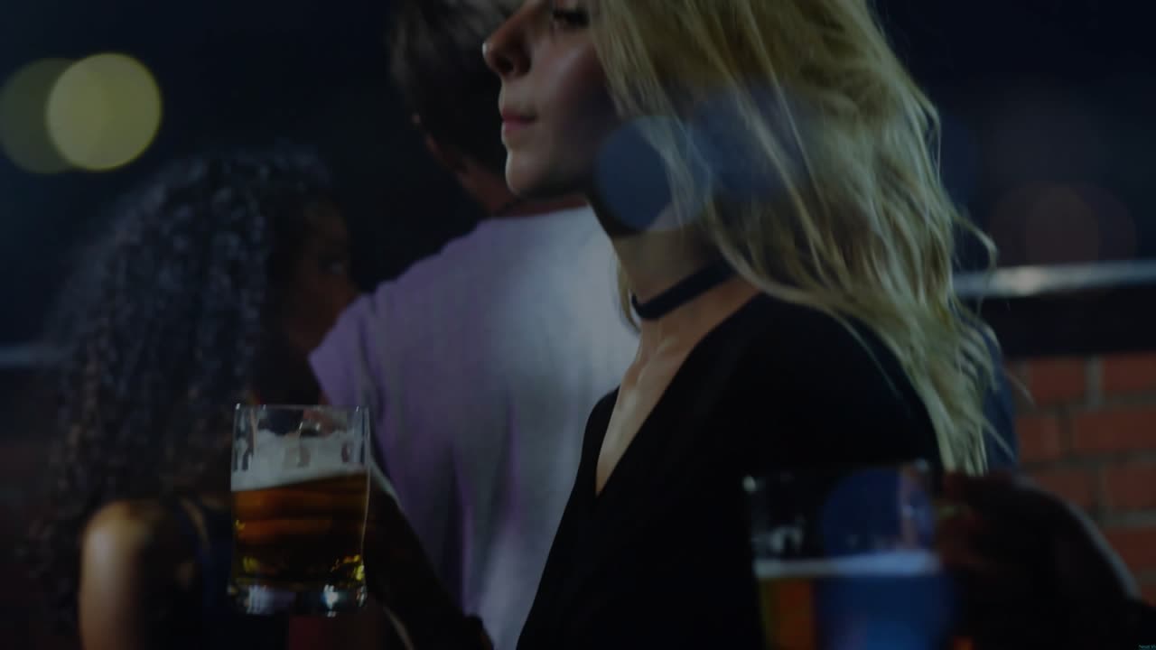 Beer mug passing toward camera revealing blonde woman straightening posture, sipping beer in bar