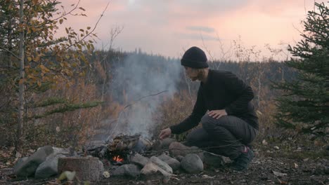 woman in black beanie and sweatshirt making campfire at alaska mountain campground during sunset - medium shot, slow motion