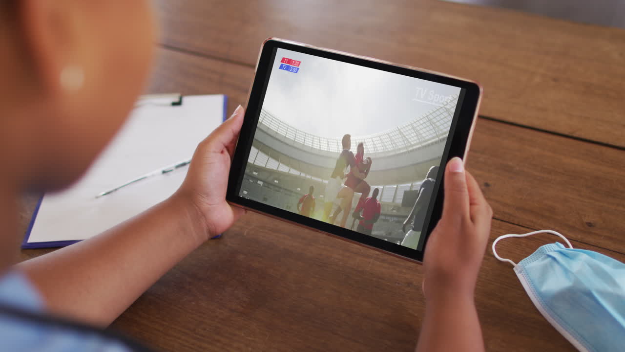 태블릿에서 럭비 경기를 시청하는 얼굴 마스크와 함께 테이블에 앉아있는 여성의 복합