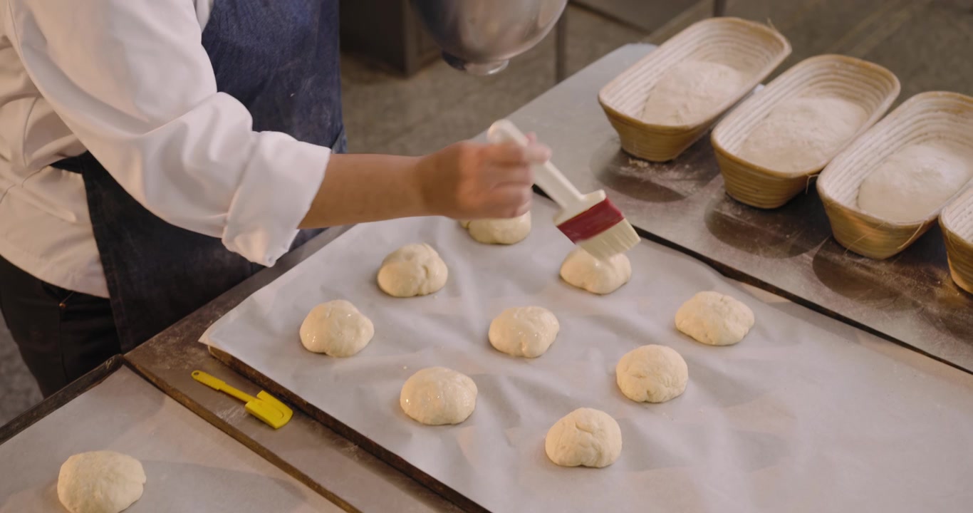 Adult African American baker after placing dough rounds brushing egg wash for proofing at bakery