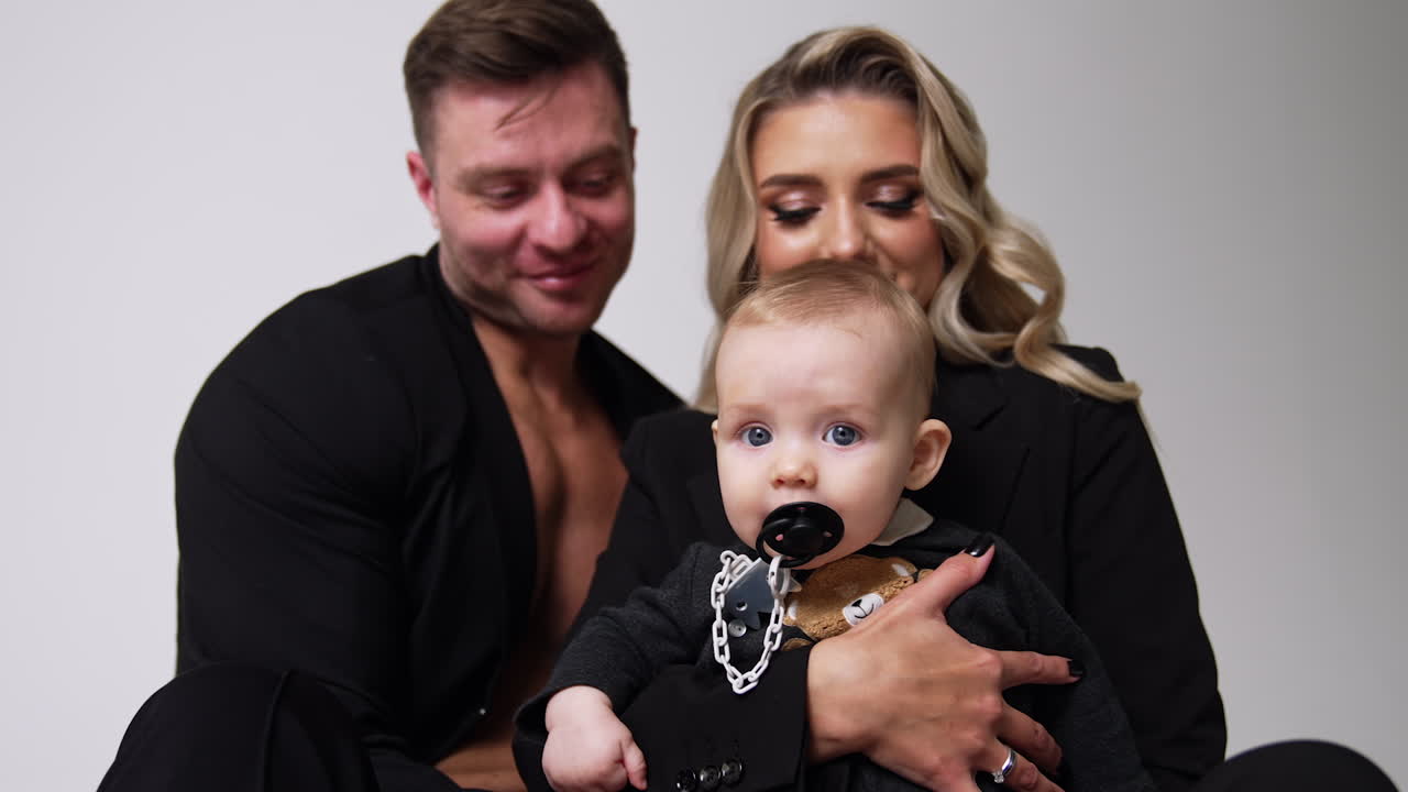 Caucasian parents wearing black jackets hold the baby with pacifier in mouth. Portrait of happy family together.
