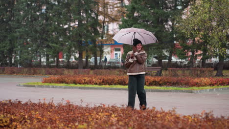 Young girl holding umbrella, wearing knit cap, brown shearling jacket, black trousers, happily spinning umbrella in light snowfall, enjoying winter day, playful and carefree in cold weather