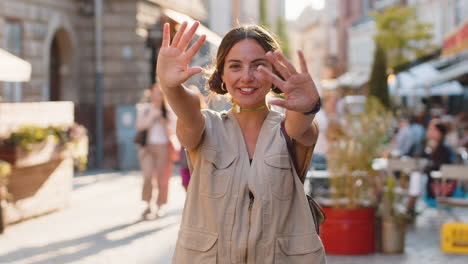 young woman smiling friendly at camera, waving hands hello, hi, greeting or goodbye on city street