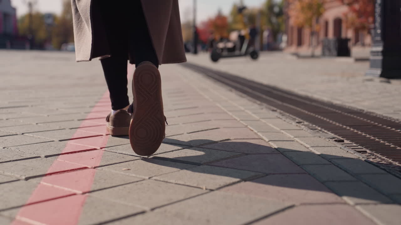 Gros plan en contre-plongée sur des pas sur le trottoir. Personne en long manteau marchant le long d'une ligne de guidage tactile rouge à côté des rails du tramway, un pigeon à proximité. Lumière du soleil d'automne projetant une ombre allongée sur les pavés texturés et le métal.