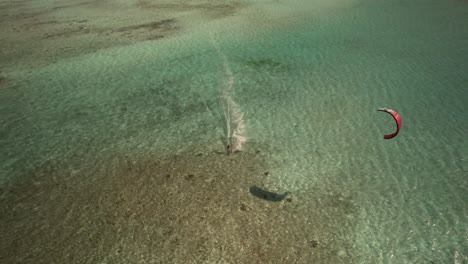 Kiteboarder-gliding-on-turquoise-waters-with-vibrant-red-kite-seen-from-above