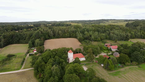 Small-hill-in-Glaznoty-covered-with-trees,-and-meadow-with-a-small-church-with-a-red-tile-roof-in-the-middle-and-dense-forest