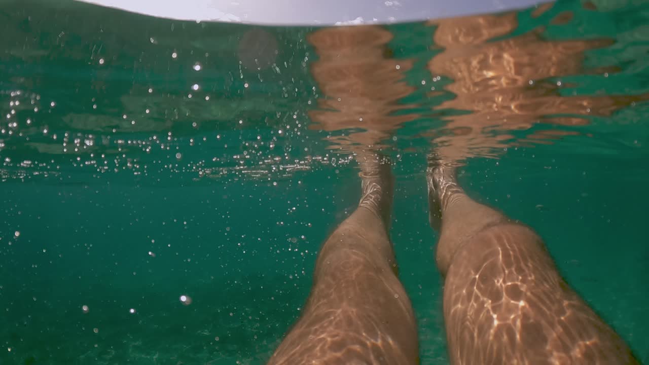 Vista subacquea in prima persona di gambe di uomo che galleggiano nell'acqua limpida e trasparente del mare con la linea di galleggiamento e la costa sullo sfondo, slow motion