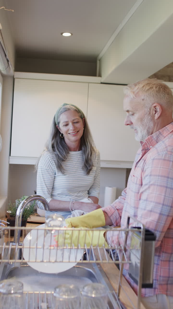 Vídeo vertical de casal caucasiano idoso feliz lavando louça na cozinha, em câmera lenta.