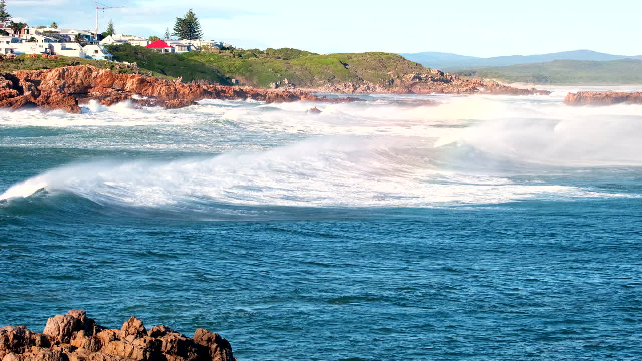 Wind blows misty sea spray over crest of breaking wave to create rainbow colors