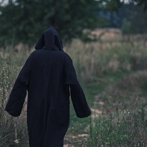 backside portrait of death in nature. dark figure in black costume out stretches its arms and goes away in the field in the evening. scary nightmare ghost outdoors.