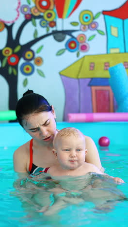brunette woman holds a cute plump baby boy in the swimming pool. coach talks to a kid who wants to cry. vertical video.