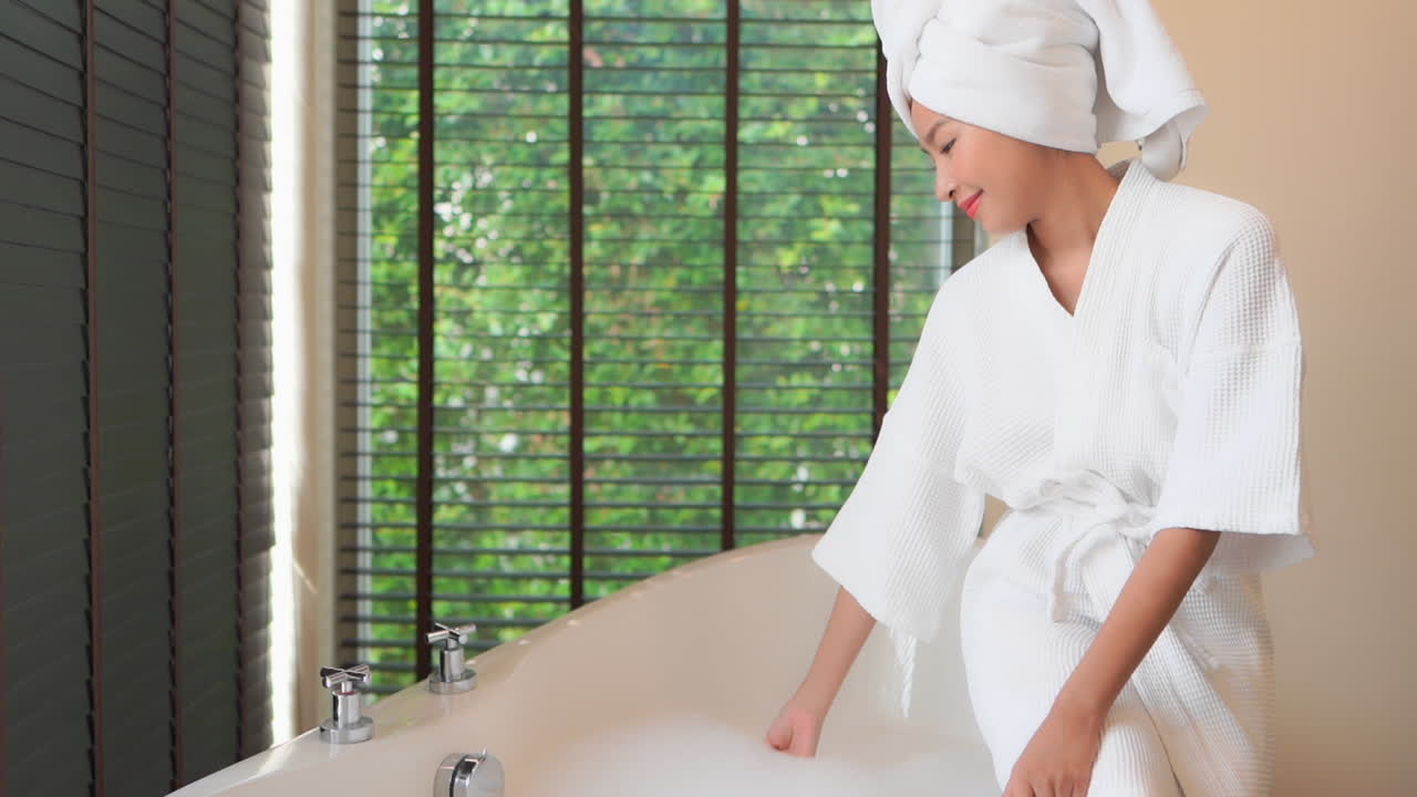 A young woman in a bathrobe and hair up in a towel turns off the water filling her bubble bath