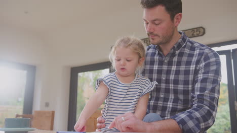 Father-at-home-at-kitchen-counter-helping-young-daughter-to-draw-picture-in-book---shot-in-slow-motion