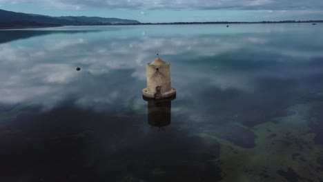 Old-Spanish-windmill-fast-orbit-in-the-lagoon-at-the-island-town-Orbetello-close-to-Monte-Argentario-and-the-Maremma-Park-in-Tuscany,-Italy,-with-blue-sky-and-calm-blue-water