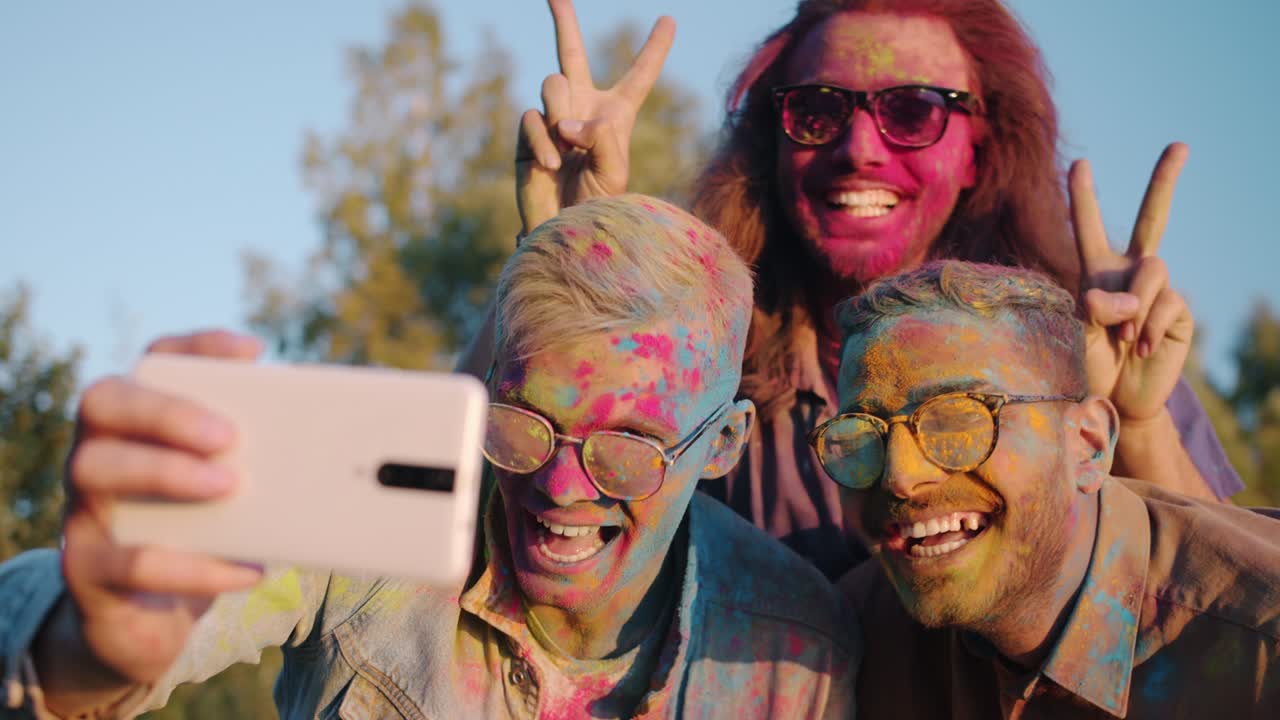 estudiantes varones felices disfrutando del festival de holi tomando selfies con caras coloridas usando la cámara del teléfono inteligente