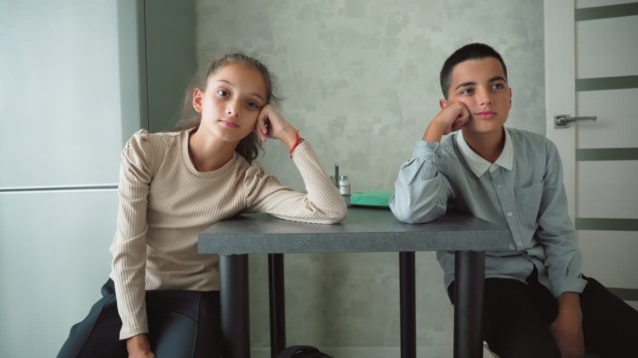 Two schoolchildren sitting quietly at kitchen table, appearing bored and thoughtful, surrounded by books and school items, possibly reflecting, waiting, or disengaged during home learning session