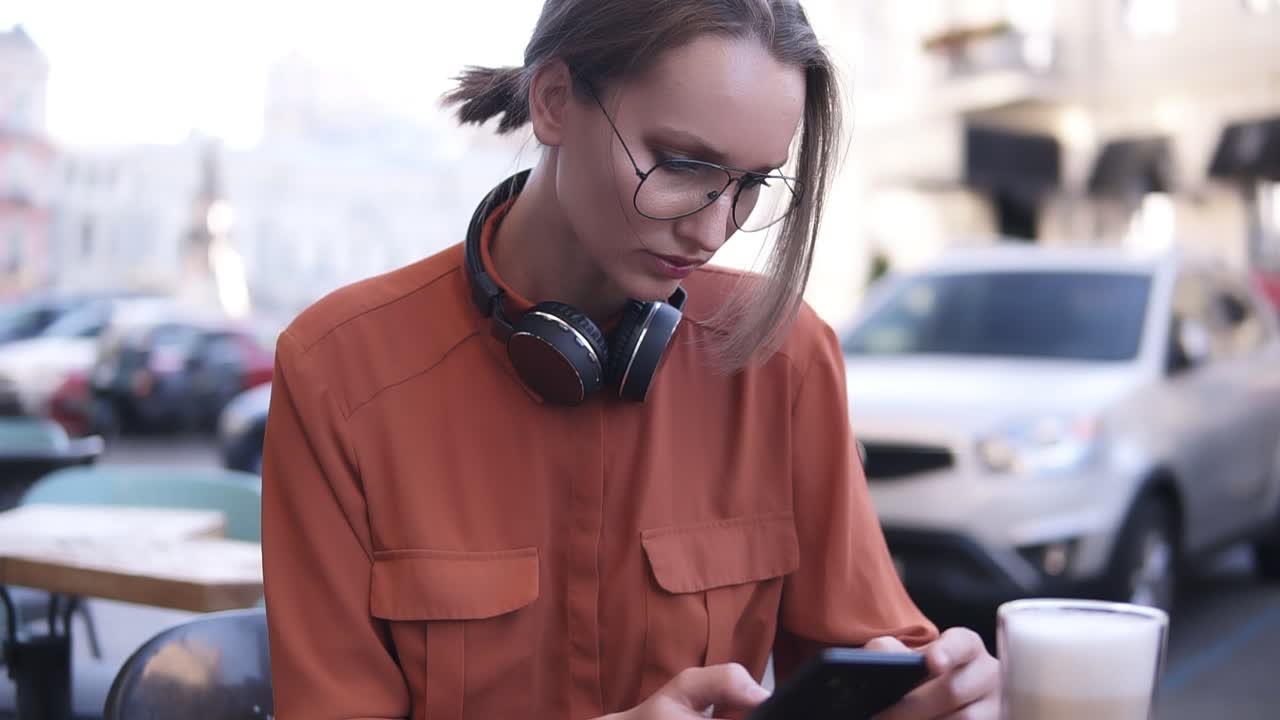 y una atractiva chica rubia con el cabello en un manojo y gafas está sentada en un café en la calle, junto a ella en la mesa de madera