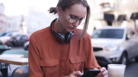 and attractive blonde girl with hair in a bundle and glasses is sitting in a cafe on the street, next to her on the wooden table