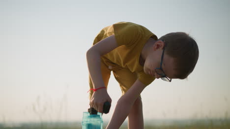 young boy wearing a yellow shirt and shorts, with glasses, bends over while holding a plastic container outdoors. the scene captures the child in a grassy field under a clear sky