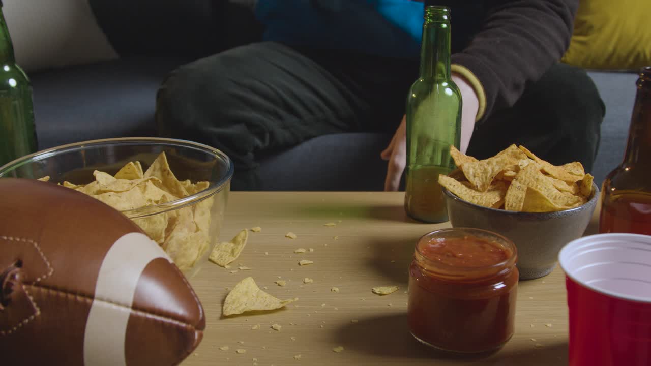 persona en casa viendo un partido de fútbol americano en la televisión con bebidas, bocadillos y pelota 1