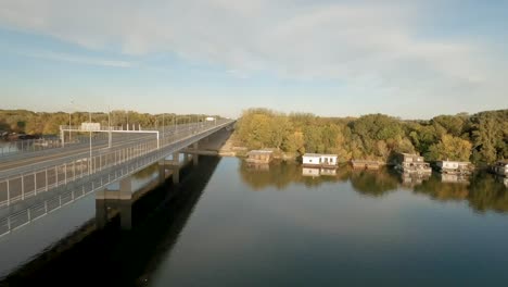 Coloridas-Casas-Flotantes-Atracadas-En-Un-Río-Tranquilo,-Con-Un-Puente-Que-Transporta-Autos-Cruzando-En-La-Distancia