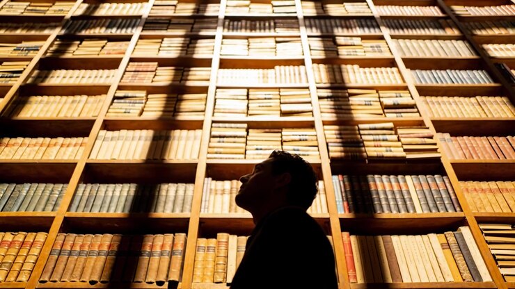 Luce calda cangiante, uomo con la giacca che inclina la testa scrutando gli scaffali in biblioteca, scala, ombre.