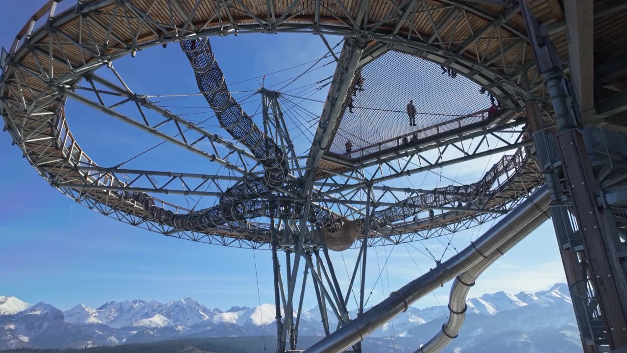 SkyWalk Serce Poronina in Male Ciche, Polen, mit Familien auf dem erhöhten Aussichtssteg, Rutsche und Netzbrücke über einer verschneiten Tatra-Berglandschaft an einem sonnigen Wintertag.