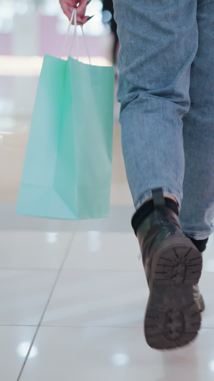 vista inferior del cuerpo de una persona caminando en azulejos elegantes del centro comercial mientras lleva dos bolsas de compras, con pantalones vaqueros y botas negras, con otros compradores moviéndose en el fondo en un entorno moderno del centro comercial