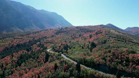 Aerial-over-forestland-showcasing-vibrant-fall-colors,-close-to-Kyhv-Peak-in-Utah,-with-a-winding-road-and-impressive-mountain-range-on-a-clear-day