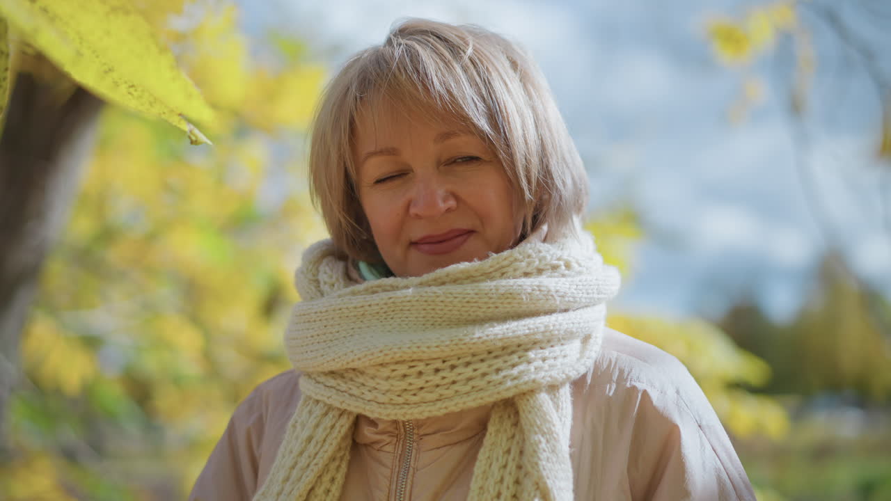 elegant woman with soft smile standing outdoors under golden autumn leaves swaying in gentle wind, dressed in light jacket and thick knitted scarf, surrounded by peaceful seasonal landscape
